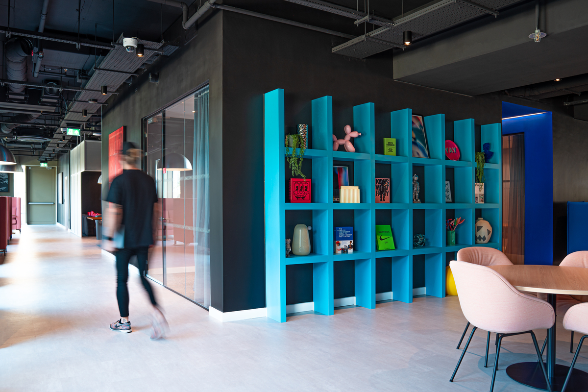 Modern office interior with a person walking past colorful shelves displaying various decorative items. The room features a black ceiling, pink chairs, and a round table, creating a vibrant and contemporary atmosphere.
